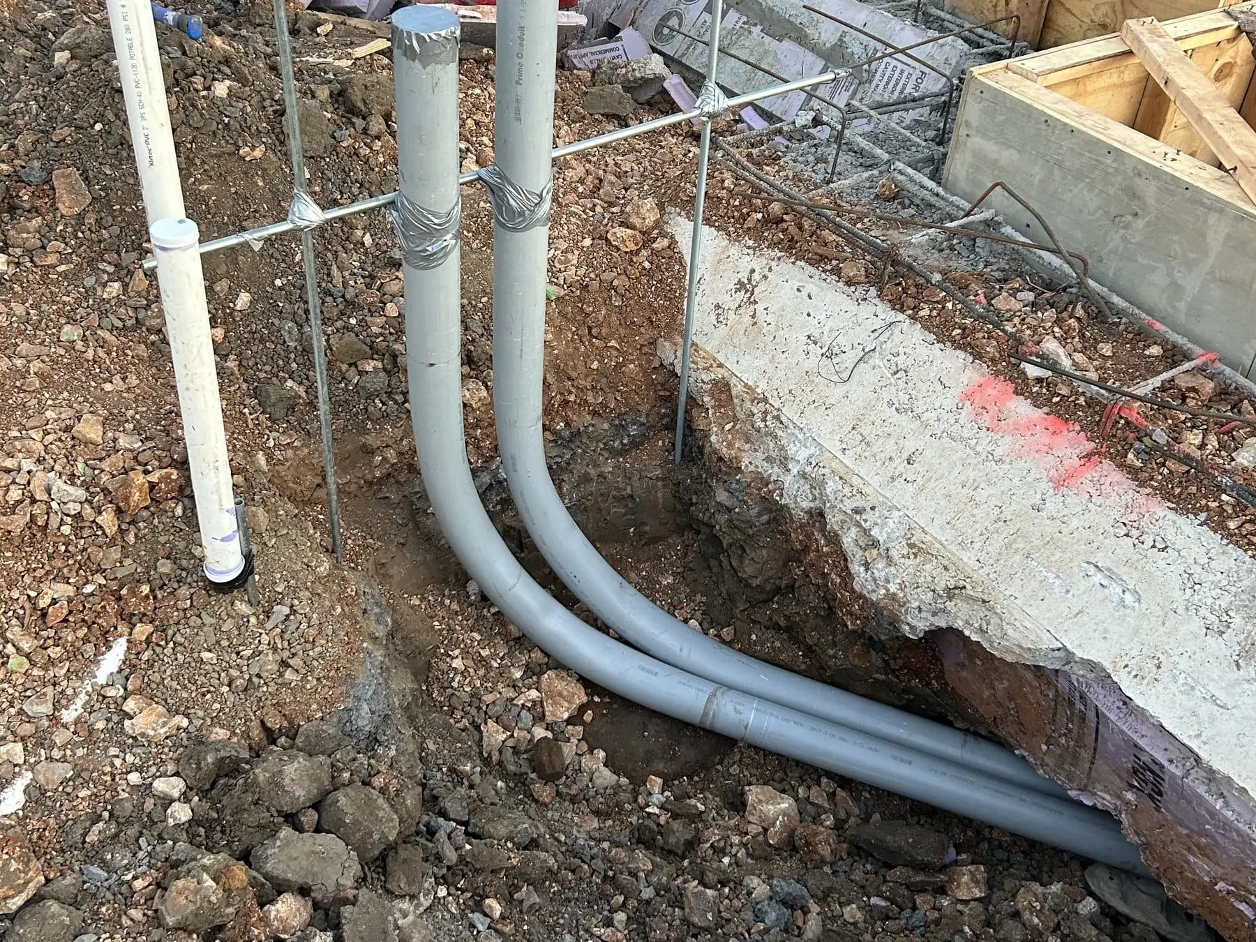 Conduit runs laid in an open trench alongside a foundation in red clay